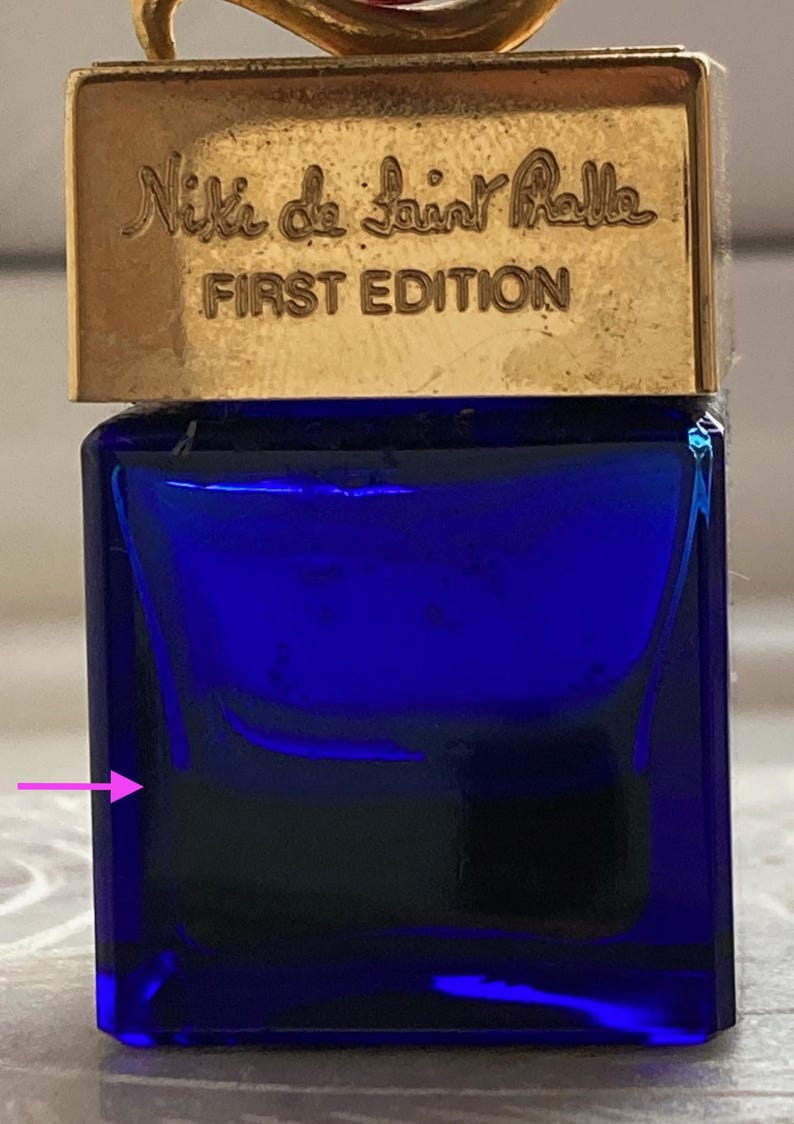 Puede incluir: Una peque&ntilde;a botella de vidrio azul con tapa dorada. La botella tiene el texto "Niki de Saint Phalle FIRST EDITION" en la parte superior.