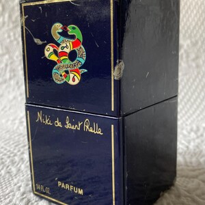 Puede incluir: Una caja azul con ribetes dorados y un dise&ntilde;o de serpiente en la parte superior. La caja dice "Niki de Saint Phalle" y "Parfum" en la parte delantera. La caja es de 1/4 fl oz o 7 ml.