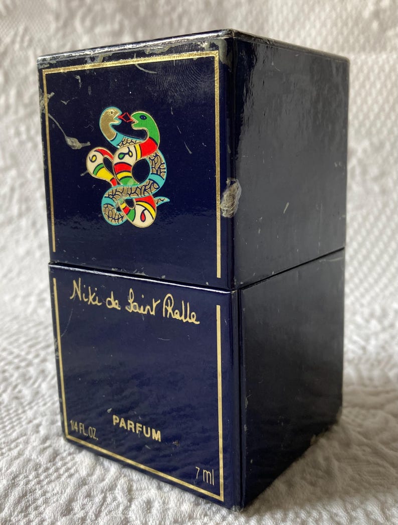 Puede incluir: Una caja azul con ribetes dorados y un dise&ntilde;o de serpiente en la parte superior. La caja dice "Niki de Saint Phalle" y "Parfum" en la parte delantera. La caja es de 1/4 fl oz o 7 ml.