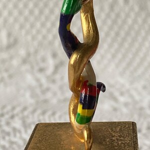 Puede incluir: Una escultura de metal dorado de una serpiente con una cola rayada de colores. La serpiente est&aacute; enroscada y se encuentra sobre una base cuadrada.