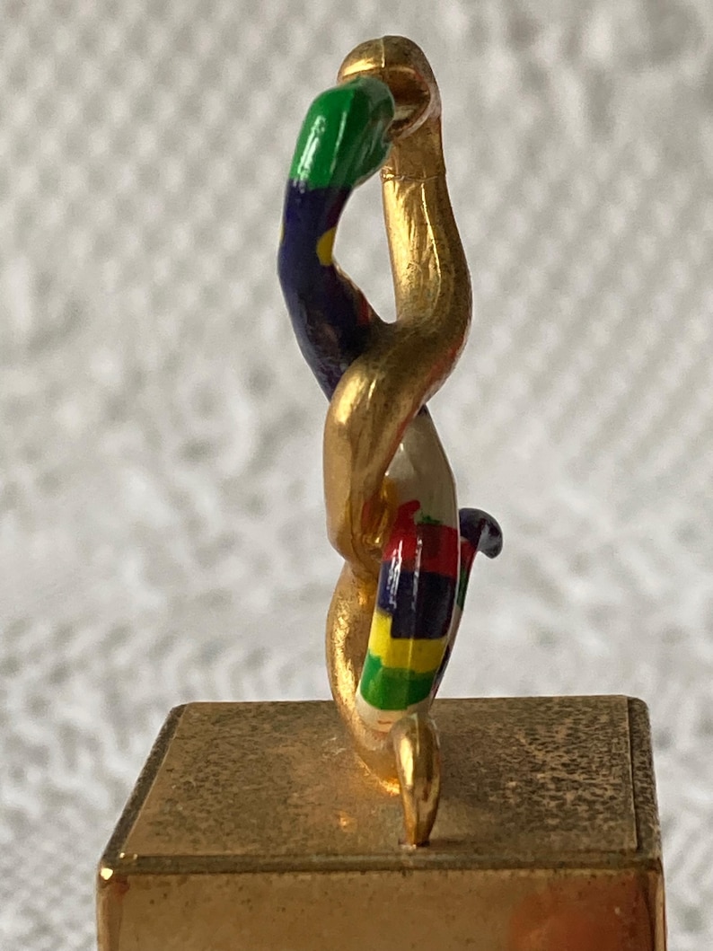 Puede incluir: Una escultura de metal dorado de una serpiente con una cola rayada de colores. La serpiente est&aacute; enroscada y se encuentra sobre una base cuadrada.