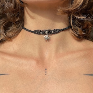 May include: A black choker necklace with a silver sun charm. The necklace is made of a thin, braided cord with small silver beads.