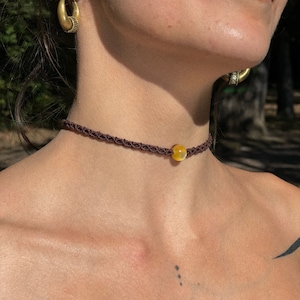 Pode incluir: Um colar gargantilha de macramê marrom com uma única conta de pedra preciosa amarela. O colar é usado ao redor do pescoço. Brincos de argola dourados são visíveis ao fundo. A joia é de estilo boêmio.