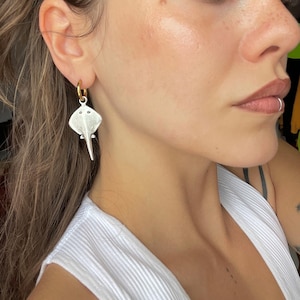 Puede incluir: Pendientes plateados con forma de raya, con aros dorados. Los pendientes presentan un diseño detallado de una raya, con una superficie texturizada y pequeños detalles de ojos. Los pendientes se llevan en la oreja.
