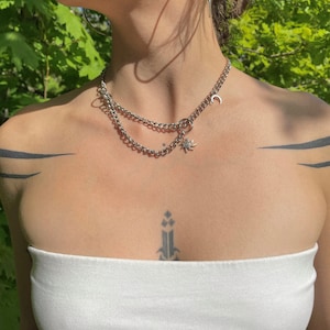 Może przedstawiać: Srebrny, warstwowy naszyjnik z wisiorkiem w kształcie półksiężyca i słońca. Naszyjnik ma podwójny łańcuszek i mały pierścień łączący warstwy. Biżuteria jest noszona na białym topie bez ramiączek.