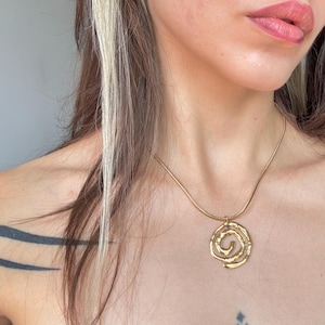 Könnte beinhalten: Eine goldfarbene Halskette mit einem runden Anhänger in einem texturierten, unregelmäßigen Spiraldesign. Der Anhänger hängt an einer glatten, dünnen Schlangenkette, die auf dem Schlüsselbein aufliegt. Die Metalloberfläche hat ein mattes, gehämmertes Finish.