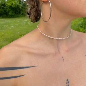 以下が含まれることがあります： 小さな白いビーズが付いたシルバーのチョーカーネックレス。ネックレスは肩にタトゥーのある人が着用しています。