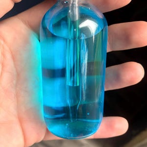 May include: A clear glass bottle filled with bright blue liquid. The bottle has a black cap and a glass dropper. The liquid is a vibrant shade of blue, and the bottle is held in a hand.