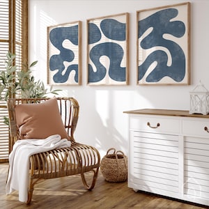 Coastal navy blue wall art set hanging in a sunlit room next to a rattan chair and a white dresser.