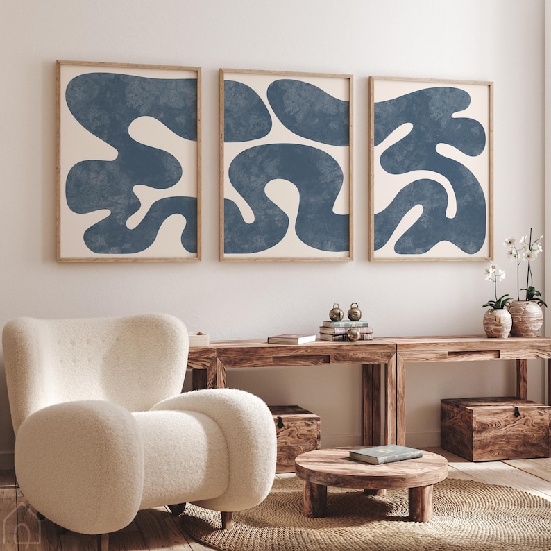 Set of 3 navy blue minimalist abstract wall art prints with organic shapes in wood frames above a white bouclé chair.