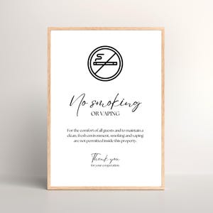 Puede incluir: Un letrero enmarcado con fondo blanco y un símbolo negro de "prohibido fumar". El texto dice "No smoking or vaping" e incluye un mensaje sobre el mantenimiento de un ambiente limpio. El letrero está en un marco de madera clara.