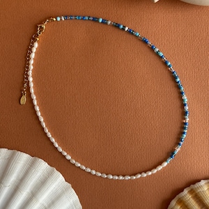 Puede incluir: Un delicado collar con una combinación de perlas blancas y cuentas azules. El collar tiene un cierre dorado y se muestra sobre una superficie marrón con detalles de conchas marinas. Las cuentas azules varían en tonos y son transparentes.