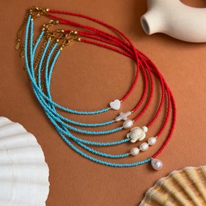 Può includere: Una collezione di collane di perline rosse e turchesi. Alcune collane presentano catene e ciondoli color oro, tra cui un cuore, una croce, una tartaruga e perle. Le collane sono esposte su una superficie marrone con conchiglie.