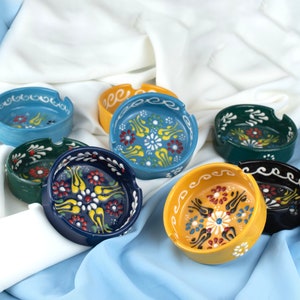 May include: A collection of ceramic ashtrays in various colors, including blue, yellow, green, and black. Each ashtray features a round shape with a decorative floral pattern in the center and along the rim. The ashtrays are displayed on a white and blue fabric.