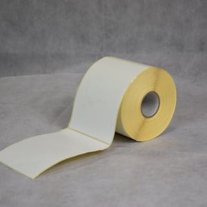 Op de afbeelding: Een rol blanco witte etiketten met een gele achterkant. De rol is gedeeltelijk afgerold, waardoor de zelfklevende kant zichtbaar is. De etiketten zitten op een rol, klaar voor gebruik. De achtergrond is neutraal grijs.
