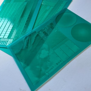 Può includere: Una pila di fogli di plastica trasparenti color teal. Ogni foglio presenta linee di griglia stampate, cerchi e altre forme geometriche. La parola "LOBOYO" è visibile su un foglio.