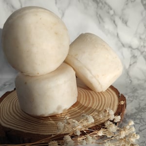 May include: Three off-white soaps, round and rectangular, are arranged on a wooden slice, with dried white flowers in the foreground. The soaps have a smooth texture. The background is a marble pattern.