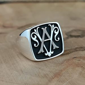 Op de afbeelding: Een zilveren zegelring met een vierkante voorkant. De voorkant heeft een zwarte achtergrond met de letter "A" in een decoratief, gestileerd lettertype. De ring heeft een gepolijste, glanzende afwerking en ligt op een houten oppervlak.