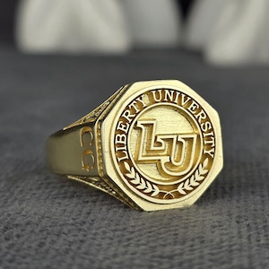 May include: A gold ring featuring the words "LIBERTY UNIVERSITY" and the letters "LU". The ring has an octagonal face. The background is a plain grey colour.