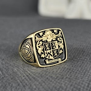 May include: A gold-colored signet ring with a black enamel face. The ring features a family crest with a helmet, shield, and the name "Mahoney". The ring has a Celtic knot design on the side.