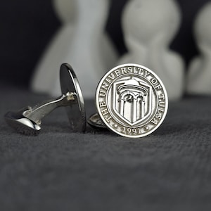 Könnte beinhalten: Silberne Manschettenknöpfe mit dem Siegel der University of Tulsa. Die runden Manschettenknöpfe zeigen ein detailliertes Emblem und das Jahr 1991. Der Hintergrund ist dunkelgrau, mit verschwommenen weißen Dekorfiguren.