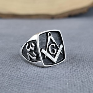 May include: Silver-toned ring with a square face featuring a Masonic symbol, including a compass, square, and the letter "G". The number "32" is on the side of the ring. The ring is set against a gray background.