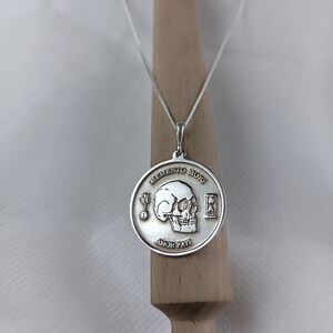 May include: Silver necklace with a round pendant featuring a skull, an hourglass, and the words "Memento Mori" and "Amor Fati".