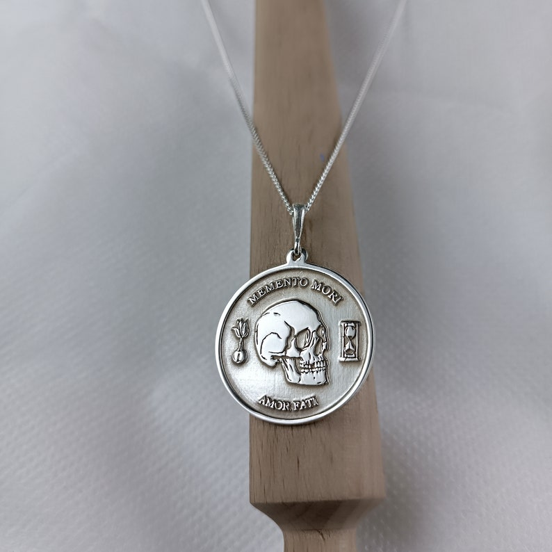 May include: Silver necklace with a round pendant featuring a skull, an hourglass, and the words "Memento Mori" and "Amor Fati".