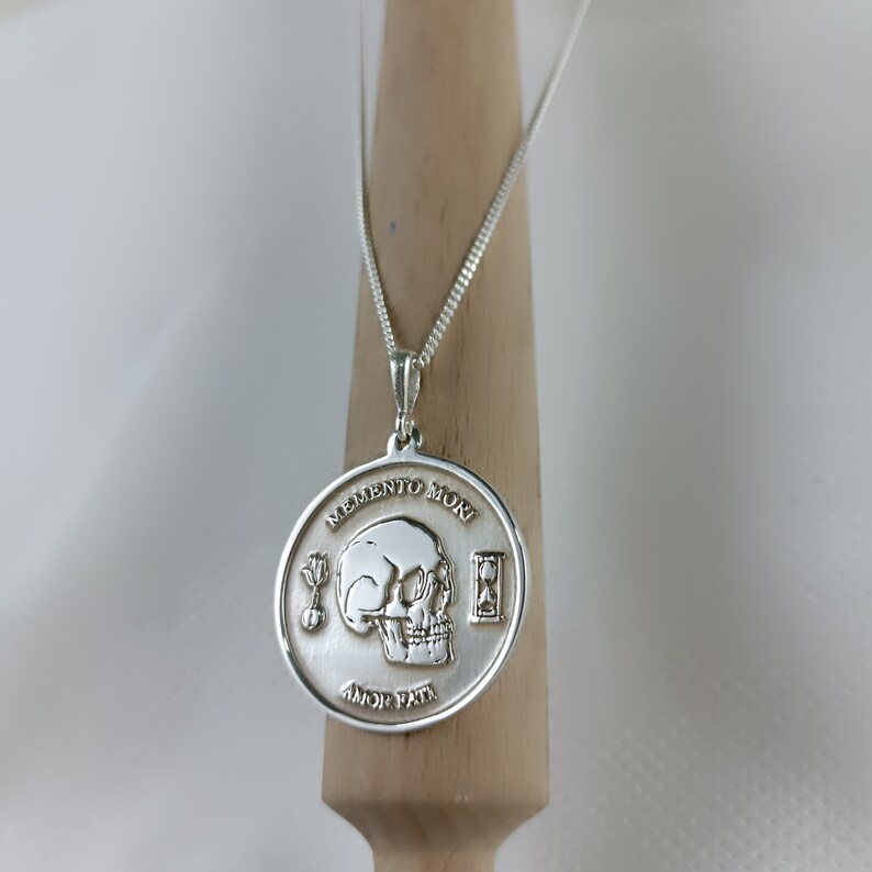 May include: Silver necklace with a round pendant featuring a skull, an hourglass, and the words "Memento Mori" and "Amor Fati".