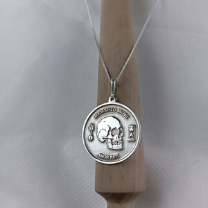 May include: A silver necklace with a round pendant featuring a skull, an hourglass, and the words "Memento Mori" and "Amor Fati".