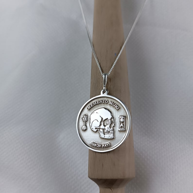 May include: A silver necklace with a round pendant featuring a skull, an hourglass, and the words "Memento Mori" and "Amor Fati".