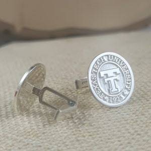Könnte beinhalten: Silberne Manschettenknöpfe mit einem runden Design, das das Logo der Texas Tech University und das Jahr 1923 zeigt.
