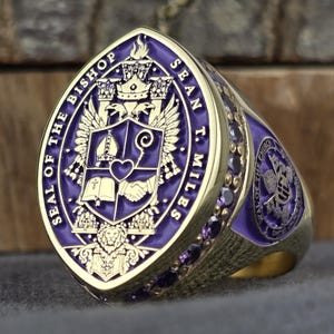 Könnte beinhalten: Ein goldener Ring mit einer großen ovalen Front und violetter Emaille-Einlage. Die Mitte zeigt ein Wappen mit Mitra, Krummstab, Herz, Buch und Händedruck. Text: SEAL OF THE BISHOP SEAN T. MILES. Die Seiten sind mit violetten Steinen besetzt.