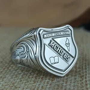 Può includere: Un anello d'argento con un designo a forma di scudo. Lo scudo presenta il testo "MCHS 85", un libro aperto e una torcia. Il testo "LABOR OMNIA VINCIT" è inciso sopra lo scudo.