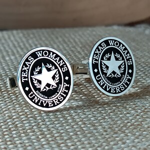 Könnte beinhalten: Ein Paar silberne Manschettenknöpfe mit schwarzem Emaille. Jeder Manschettenknopf zeigt das Logo der Texas Woman's University, das einen Stern, einen Lorbeerkranz und den Text "Texas Woman's University" und "Scientia Lumen Vitae" enthält.
