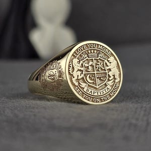 Puede incluir: Un anillo de sello dorado con un escudo grabado. El escudo presenta leones, una corona y las palabras "I love you more, see you on the moon, Baptista". El anillo tiene un acabado pulido y un diseño detallado.
