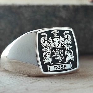 May include: A silver signet ring with a black enamel crest featuring a heraldic lion and the word "ROSS" in white.