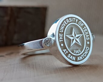 May include: Silver ring with a round, engraved design featuring a star and the text "The University of Texas at San Antonio 1969".