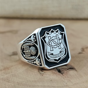 Puede incluir: Anillo de plata con una cara rectangular con un diseño de escudo en blanco y negro y el texto "A Posse Ad Esse". El anillo tiene un diseño detallado en los laterales, incluyendo un libro y una corona de laurel.