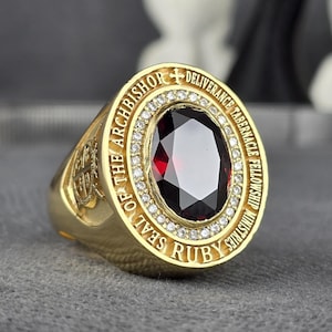 Puede incluir: Un anillo dorado con una gran piedra preciosa ovalada de color rojo oscuro en el centro. El anillo está inscrito con texto alrededor de la piedra preciosa y presenta un halo de pequeñas piedras transparentes. Es probable que el anillo sea un artículo religioso o conmemorativo.