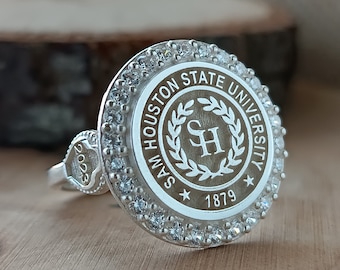 Puede incluir: Un anillo de plata con un centro redondo, blanco y marrón que presenta el logotipo de la Universidad Estatal Sam Houston y el año 1879. El anillo está decorado con pequeñas piedras transparentes alrededor del borde.