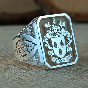Op de afbeelding: Een zilveren ring met een bruine rechthoekige inleg met een wapenschild met drie druppelvormige objecten en de tekst "Cleary".