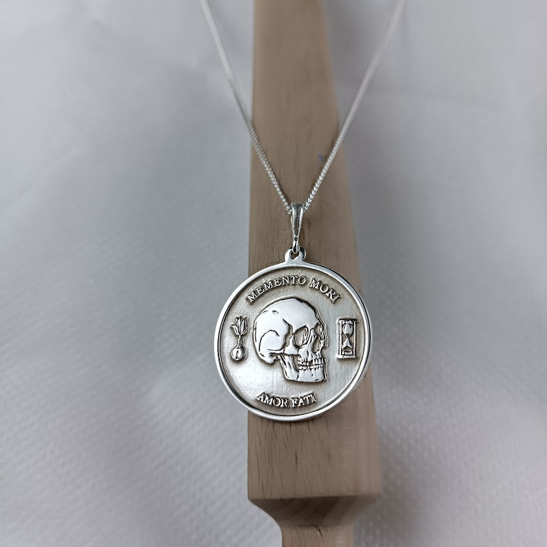 May include: Silver necklace with a round pendant featuring a skull, an hourglass, and the text "Memento Mori Amor Fati".