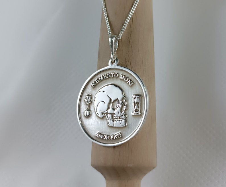May include: Silver pendant necklace with a skull and hourglass design. The pendant features the Latin phrase "Memento Mori" and "Amor Fati" inscribed around the edge.
