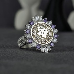May include: A silver ring featuring the Tarleton State University logo. The ring has a circular face with the university emblem, surrounded by purple and clear gemstones. The band is also adorned with clear gemstones.