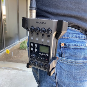 May include: A gray Zoom PodTrak P4 portable audio recorder with a microphone attached. The recorder is clipped to a person's belt loop.