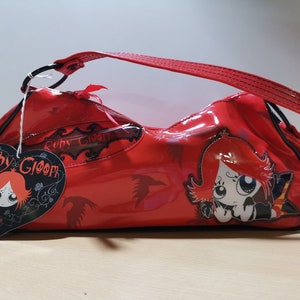 Könnte beinhalten: Eine glänzende, rote Handtasche mit geschwungener Form und passendem Henkel. Die Tasche zeigt eine Cartoon-Grafik eines Mädchens mit roten Haaren und dem Text "Ruby Gloom". Ein herzförmiges Etikett mit derselben Figur ist angebracht.