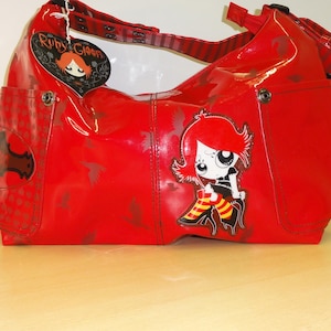 Op de afbeelding: Een rode handtas met een glanzende afwerking, met een cartoonkarakterafbeelding op de voorkant. De tas heeft een schouderriem en een hartvormig label met de tekst "Ruby Gloom". De tas heeft een zijvak.