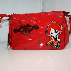 Könnte beinhalten: Eine rote Umhängetasche aus Lackleder mit einer Cartoon-Figur. Die Tasche hat einen schwarzen Riemen, einen Reißverschluss und ein dekoratives Etikett mit der Aufschrift "Ruby Gloom". Die Tasche hat ein glänzendes Finish.