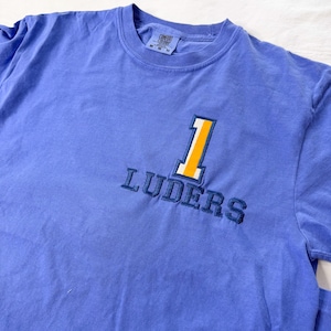 Könnte beinhalten: Ein kurzärmeliges T-Shirt in Kornblumenblau mit einer Stickerei auf der Brust. Das Motiv besteht aus einer großen gelben Eins mit weißer Umrandung über dem dunkelblauen Schriftzug LUDERS, der in Großbuchstaben gestickt ist.